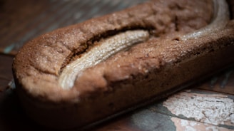 A freshly baked loaf of banana bread rests on a rustic wooden surface. The loaf has a golden-brown crust and a visible split on top, revealing a baked banana within.