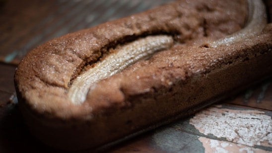 A freshly baked loaf of banana bread rests on a rustic wooden surface. The loaf has a golden-brown crust and a visible split on top, revealing a baked banana within.