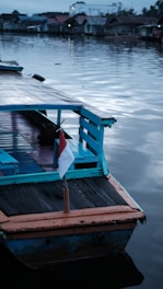 A wooden boat with blue and orange accents is anchored by a calm body of water. A small Indonesian flag is attached to the rear of the boat. In the background, there are several small houses with tin roofs lined along the waterfront. The sky and water both have a muted, grayish-blue hue, suggesting early morning or late afternoon lighting.