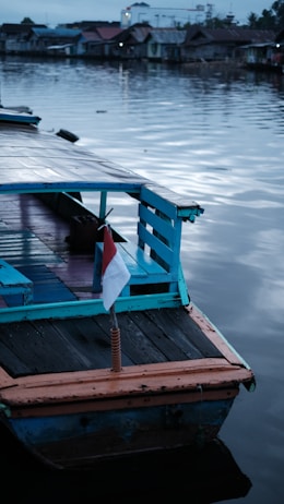 A wooden boat with blue and orange accents is anchored by a calm body of water. A small Indonesian flag is attached to the rear of the boat. In the background, there are several small houses with tin roofs lined along the waterfront. The sky and water both have a muted, grayish-blue hue, suggesting early morning or late afternoon lighting.