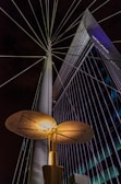 An illuminated modern streetlight with a unique, disk-shaped design is prominently featured against a backdrop of a tall, angular building with grid-like windows. Steel cables extend from the streetlight, creating a radial pattern against the night sky.