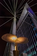 An illuminated modern streetlight with a unique, disk-shaped design is prominently featured against a backdrop of a tall, angular building with grid-like windows. Steel cables extend from the streetlight, creating a radial pattern against the night sky.