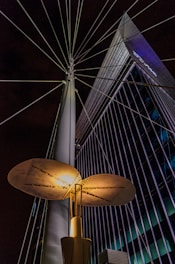 An illuminated modern streetlight with a unique, disk-shaped design is prominently featured against a backdrop of a tall, angular building with grid-like windows. Steel cables extend from the streetlight, creating a radial pattern against the night sky.