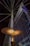 An illuminated modern streetlight with a unique, disk-shaped design is prominently featured against a backdrop of a tall, angular building with grid-like windows. Steel cables extend from the streetlight, creating a radial pattern against the night sky.