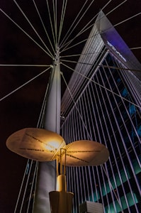 An illuminated modern streetlight with a unique, disk-shaped design is prominently featured against a backdrop of a tall, angular building with grid-like windows. Steel cables extend from the streetlight, creating a radial pattern against the night sky.