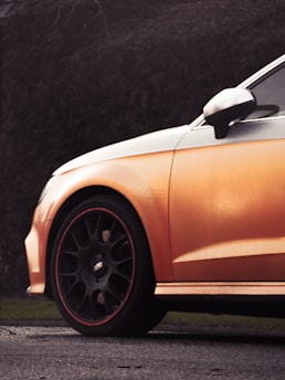 Partial view of a car with an orange body, focusing on its front side and black alloy wheel with a red rim. The background is a blurred, dark green foliage, suggesting the car is parked near a hedge.