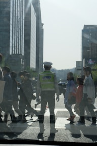 A traffic officer in a reflective vest stands in the middle of a busy crosswalk, guiding pedestrians across a city street. The background features tall modern buildings under a clear sky. People of varying appearances and ages are walking purposefully in different directions.