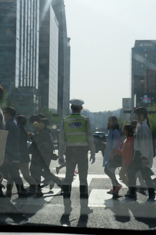A traffic officer in a reflective vest stands in the middle of a busy crosswalk, guiding pedestrians across a city street. The background features tall modern buildings under a clear sky. People of varying appearances and ages are walking purposefully in different directions.