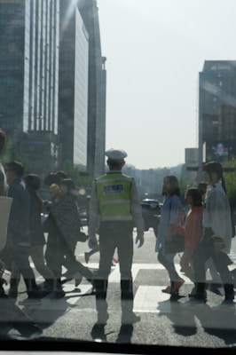 A traffic officer in a reflective vest stands in the middle of a busy crosswalk, guiding pedestrians across a city street. The background features tall modern buildings under a clear sky. People of varying appearances and ages are walking purposefully in different directions.