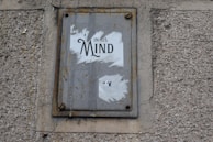 A worn and rusted metal sign is mounted on a textured concrete wall. The sign displays partially torn white paper with the word 'Mind' prominently visible, suggesting some worn-out message.
