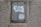 A worn and rusted metal sign is mounted on a textured concrete wall. The sign displays partially torn white paper with the word 'Mind' prominently visible, suggesting some worn-out message.