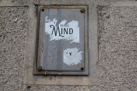 A worn and rusted metal sign is mounted on a textured concrete wall. The sign displays partially torn white paper with the word 'Mind' prominently visible, suggesting some worn-out message.