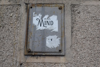 A worn and rusted metal sign is mounted on a textured concrete wall. The sign displays partially torn white paper with the word 'Mind' prominently visible, suggesting some worn-out message.
