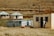 A rural scene featuring small, modest buildings with metal roofs situated on a grassy landscape. People are standing near the entrance of a whitewashed house. In the background, there are two circular structures with blue metal roofs, and cows can be seen grazing on the hilly, dry grassland.