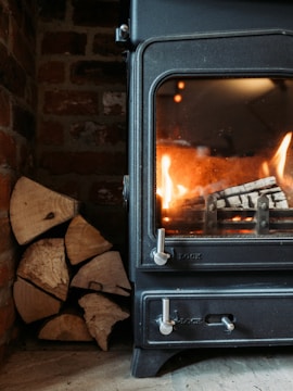 A dark metal wood-burning stove with visible flames inside is positioned against a rustic brick wall. A stack of cut logs is neatly arranged beside the stove, ready for use.