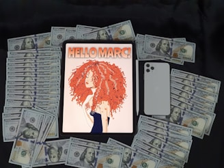 A tablet displaying an illustration of a woman with curly red hair and the text 'HELLO MARC!' on the screen. Surrounding the tablet are stacks of hundred-dollar bills. Adjacent to the tablet is a white smartphone and a stylus pen.