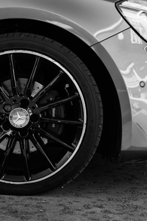 A detailed close-up of a car wheel with a prominent Mercedes-Benz logo on the hubcap. The wheel showcases sleek black spokes against a polished tire with clear treads. The body of the car is visible in the background with a smooth metallic finish.