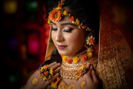 A woman adorned in elaborate bridal attire, featuring intricate jewelry and floral decorations. Her eyes are closed, suggesting a moment of introspection or serenity. The veil is embroidered with detailed patterns, and her makeup is elegantly done, emphasizing her features.
