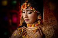 A woman adorned in elaborate bridal attire, featuring intricate jewelry and floral decorations. Her eyes are closed, suggesting a moment of introspection or serenity. The veil is embroidered with detailed patterns, and her makeup is elegantly done, emphasizing her features.