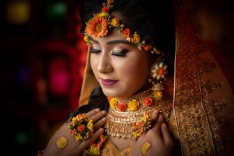 A woman adorned in elaborate bridal attire, featuring intricate jewelry and floral decorations. Her eyes are closed, suggesting a moment of introspection or serenity. The veil is embroidered with detailed patterns, and her makeup is elegantly done, emphasizing her features.