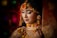 A woman adorned in elaborate bridal attire, featuring intricate jewelry and floral decorations. Her eyes are closed, suggesting a moment of introspection or serenity. The veil is embroidered with detailed patterns, and her makeup is elegantly done, emphasizing her features.