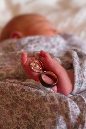 A newborn baby is wrapped in a floral-patterned blanket, with a close-up view of the baby's foot. Two rings are delicately placed around the baby's toes, one appearing to be a wedding band and the other possibly an engagement ring. The baby is lying on a soft surface, with a blurred background.