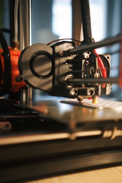 A close-up of a 3D printer in action, focusing on the extruder head as it moves over the print bed. The background is blurred, highlighting the precision machinery and components such as gears and electric wires. The scene is well-lit, emphasizing the mechanical details of the printer.