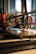 A close-up of a 3D printer in action, focusing on the extruder head as it moves over the print bed. The background is blurred, highlighting the precision machinery and components such as gears and electric wires. The scene is well-lit, emphasizing the mechanical details of the printer.