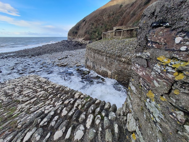 A cobblestone pathway and retaining walls made of textured stone leading towards a rocky shoreline, with waves crashing against the rocks. The landscape features rugged cliffs, partially covered with vegetation, in the background. The sky is clear with hints of clouds, contributing to a serene coastal scene.