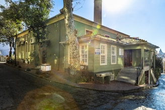 A charming green building with large windows, surrounded by trees, sits at the corner of a cobblestone street. The building has a chimney and a small staircase at the front. Sunlight is filtering through the trees, casting a beautiful flare effect and illuminating the scene with a soft glow.