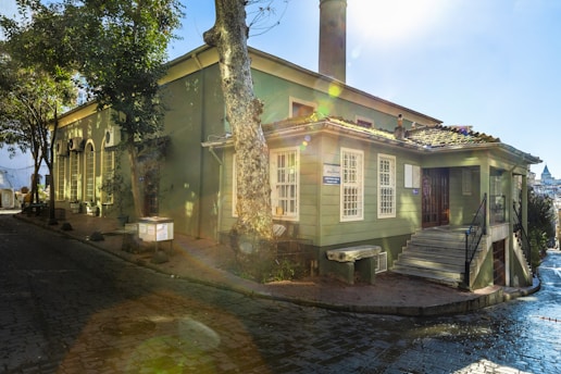 A charming green building with large windows, surrounded by trees, sits at the corner of a cobblestone street. The building has a chimney and a small staircase at the front. Sunlight is filtering through the trees, casting a beautiful flare effect and illuminating the scene with a soft glow.