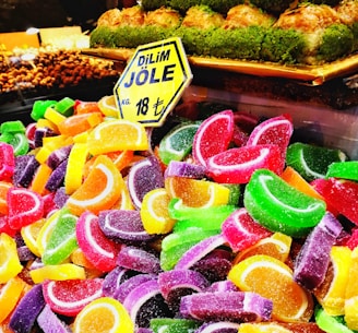 A colorful assortment of sugar-coated fruit jelly candies shaped like citrus slices is displayed in a vibrant and attractive arrangement. A sign in the background indicates the price in Turkish currency, and above it, a tray of baklava topped with green pistachio can be seen.
