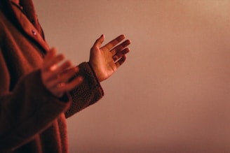 A person wearing a brown, textured jacket is holding their hands up in an open and relaxed manner. The background is a soft, neutral shade, creating a warm and calming atmosphere.