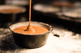 A stream of chocolate batter is being poured into a round baking pan. The background is blurred, highlighting the flow and texture of the creamy mixture.