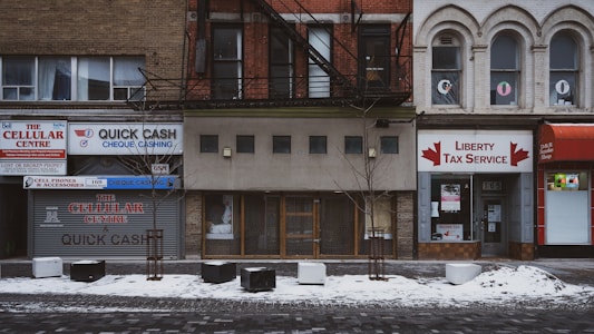 This is a street view of several storefronts on a snowy day. The businesses include a cellular center, a cheque cashing service, and a tax service. The buildings are older, with distinct brick facades and some arched windows. There are small trees planted in front of the buildings, with some snow covering the ground.