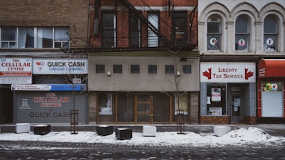 This is a street view of several storefronts on a snowy day. The businesses include a cellular center, a cheque cashing service, and a tax service. The buildings are older, with distinct brick facades and some arched windows. There are small trees planted in front of the buildings, with some snow covering the ground.