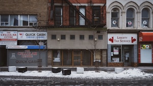 This is a street view of several storefronts on a snowy day. The businesses include a cellular center, a cheque cashing service, and a tax service. The buildings are older, with distinct brick facades and some arched windows. There are small trees planted in front of the buildings, with some snow covering the ground.