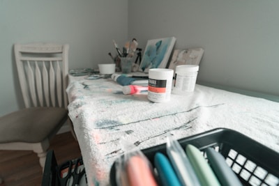 A table is covered with a cloth stained with paint, holding several art supplies including paint jars, brushes in a cup, and a tube of paint. There are paintings leaning against the wall in the background. A chair is positioned to the side, and various art tools are neatly stored in a nearby basket.