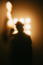 A silhouette of a person stands against a bright background with multiple circular lights. The image is heavily blurred, giving an abstract effect. The lighting creates a warm, golden glow, and the person appears to be making a gesture with their hand near their face.