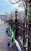 Several green electric scooters are lined up against a black wrought iron fence on a snow-dusted sidewalk. Leafless trees and classic street lamps can be seen, with people walking in the distance. A modern building is visible on the street corner, creating an urban, wintry setting.