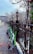 Several green electric scooters are lined up against a black wrought iron fence on a snow-dusted sidewalk. Leafless trees and classic street lamps can be seen, with people walking in the distance. A modern building is visible on the street corner, creating an urban, wintry setting.