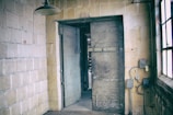 An industrial setting with a metal door slightly ajar, leading to another dimly lit room. The walls are constructed from large, weathered bricks, and there is a hanging light fixture above. A window to the right provides some natural light, and electrical wiring and a meter are mounted on the wall next to the door.