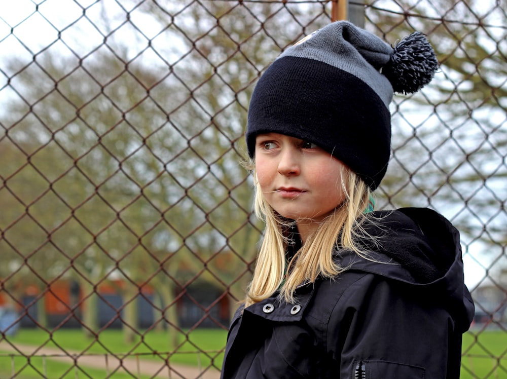 woman in black knit cap and black coat standing near gray metal fence during daytime