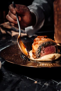person holding stainless steel fork and knife slicing meat on black ceramic plate
