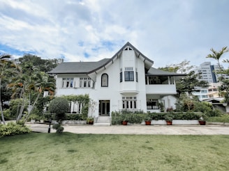 A large, elegant white house with a steep roof and multiple windows set in a lush green landscape. The foreground features a well-maintained lawn and various plants, including tall palm trees and well-trimmed bushes. The sky above is partly cloudy, adding to the serene atmosphere.