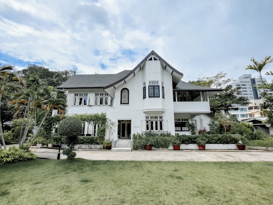 A large, elegant white house with a steep roof and multiple windows set in a lush green landscape. The foreground features a well-maintained lawn and various plants, including tall palm trees and well-trimmed bushes. The sky above is partly cloudy, adding to the serene atmosphere.