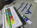 A set of acrylic paint tubes is arranged neatly in a white plastic tray, with various colors labeled on each tube. In the background, there is a grey fabric or t-shirt featuring blue calligraphy or script. A single tube of paint is placed beside the tray.