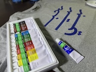 A set of acrylic paint tubes is arranged neatly in a white plastic tray, with various colors labeled on each tube. In the background, there is a grey fabric or t-shirt featuring blue calligraphy or script. A single tube of paint is placed beside the tray.