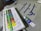 A set of acrylic paint tubes is arranged neatly in a white plastic tray, with various colors labeled on each tube. In the background, there is a grey fabric or t-shirt featuring blue calligraphy or script. A single tube of paint is placed beside the tray.