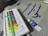 A set of acrylic paint tubes is arranged neatly in a white plastic tray, with various colors labeled on each tube. In the background, there is a grey fabric or t-shirt featuring blue calligraphy or script. A single tube of paint is placed beside the tray.